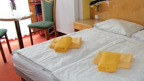 A hotel room with two beds, each with white linens and yellow towels. There is a wooden headboard, a bedside table, and a chair with a cushion.