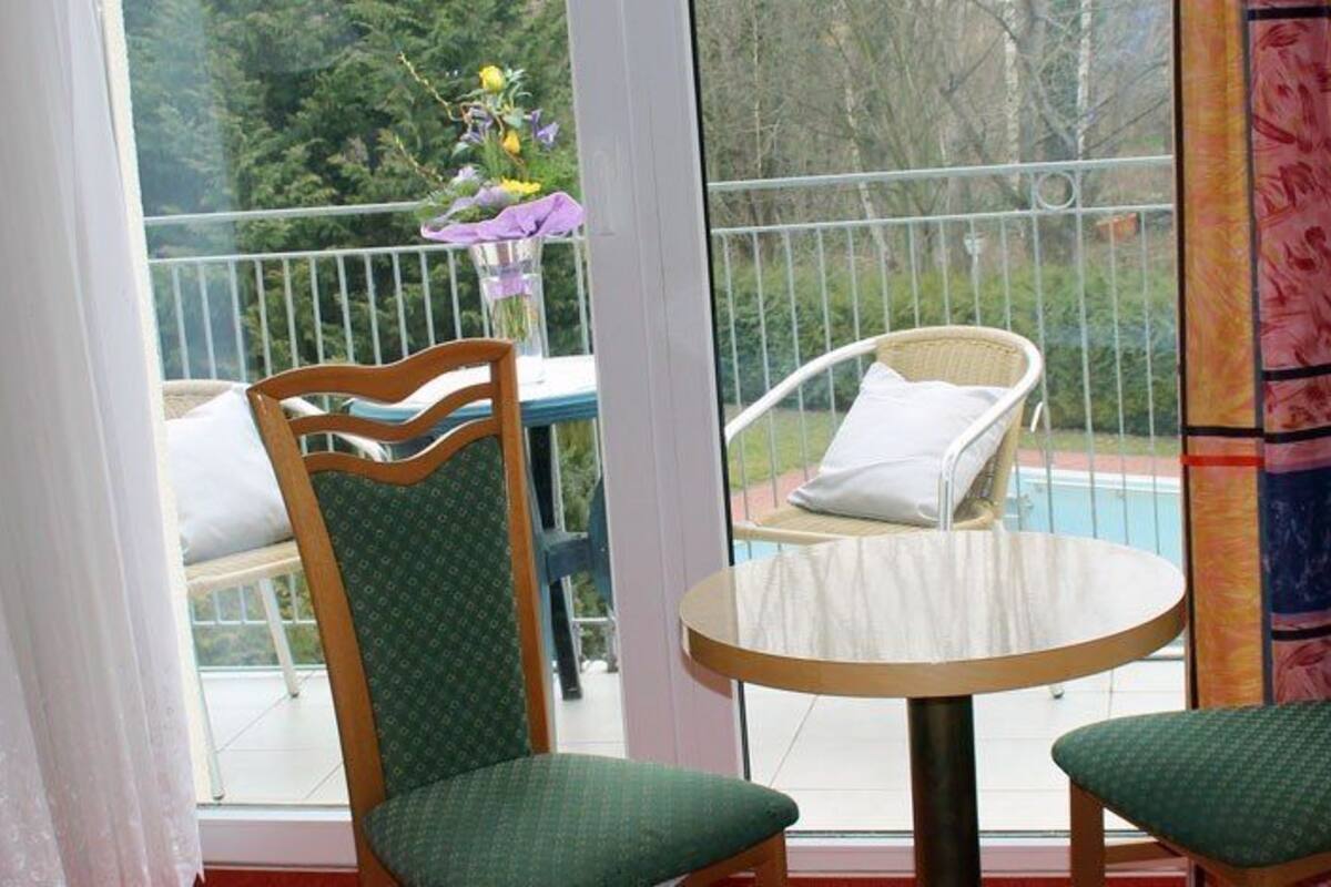 Ein Balkon mit zwei Stühlen und einem Tisch, Blick auf einen Swimmingpool und ein Fenster mit Vorhängen.