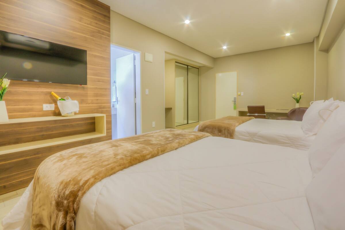 A hotel room with two beds, a TV mounted on the wall, a small table with a vase of flowers, and a sliding door leading to a bathroom.