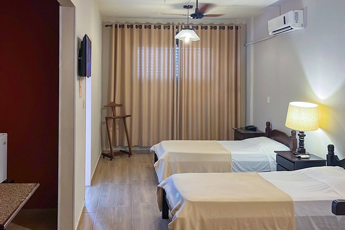 A hotel room with two beds, a desk, a lamp, a ceiling fan, and a window with curtains.