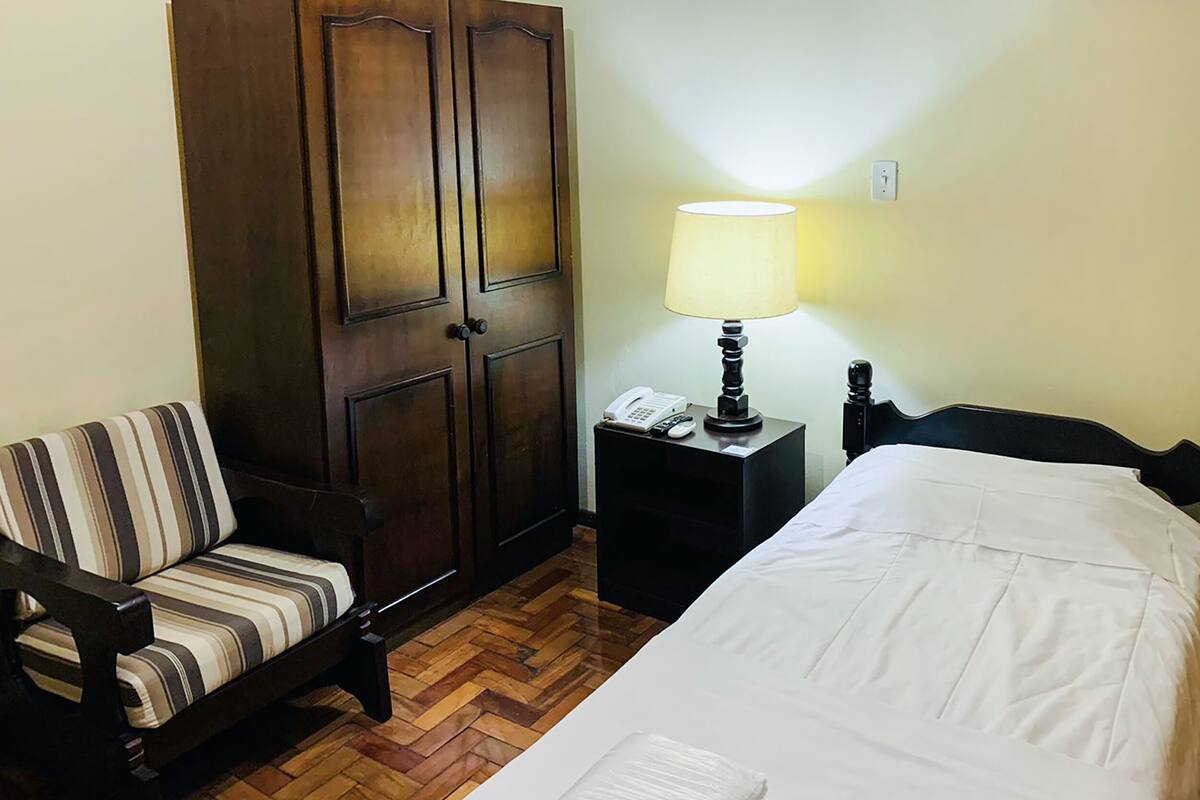 A hotel room with a bed, a chair, a wardrobe, a nightstand with a lamp, and a telephone.