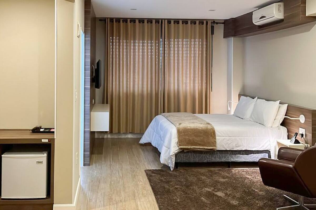 A hotel room with a bed, bedside table, chair, and a small refrigerator.