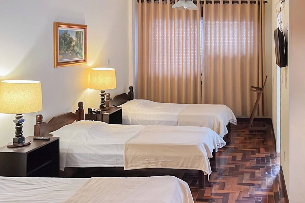 A hotel room with three single beds, a desk with a lamp, a painting on the wall, and a television mounted on the wall.