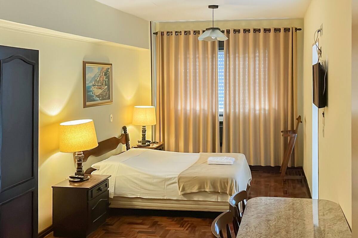 A hotel room with a bed, two bedside lamps, a painting on the wall, a wooden floor, and a curtain.