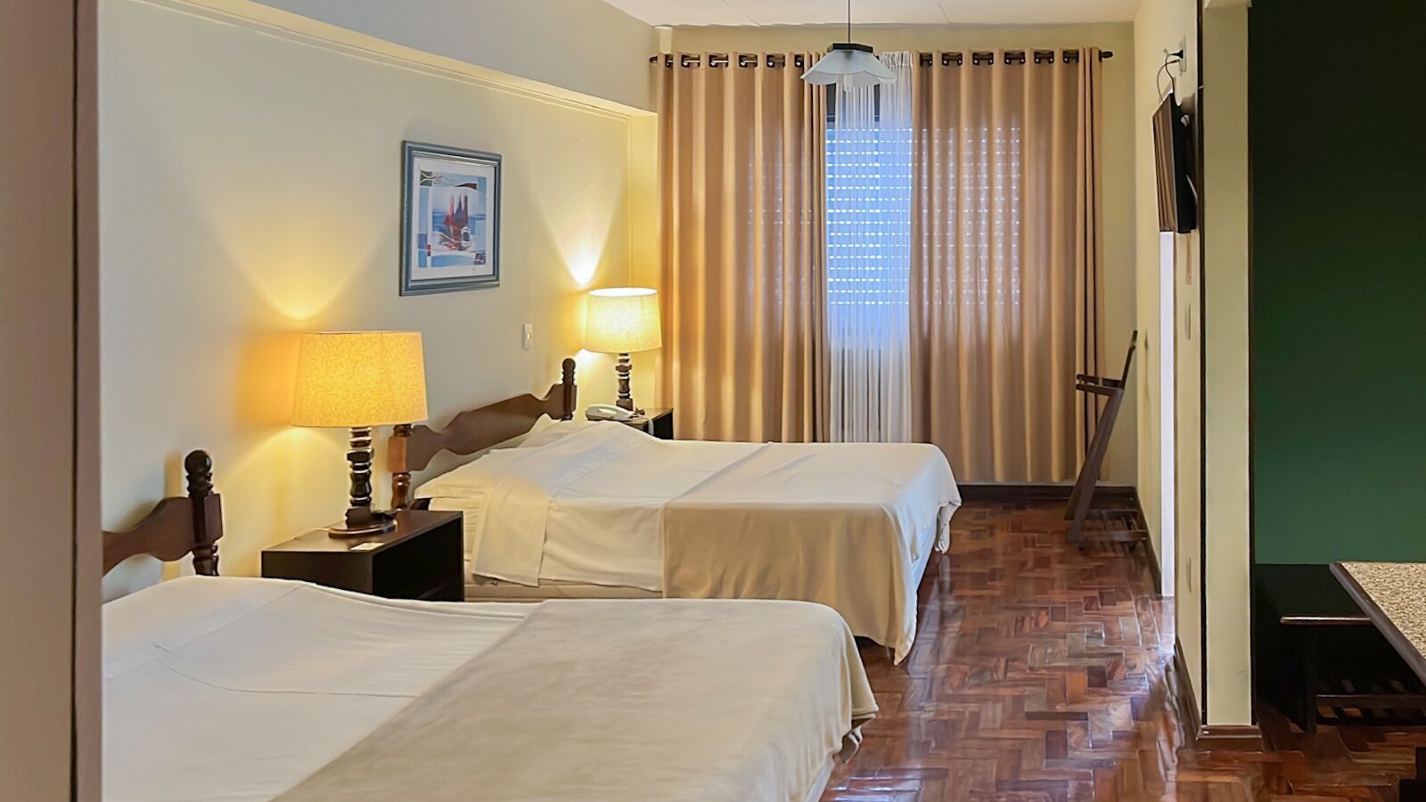 Una habitación de hotel con dos camas, un cabecero de madera, una mesita de noche con lámpara, un cuadro enmarcado en la pared y una ventana con cortinas.