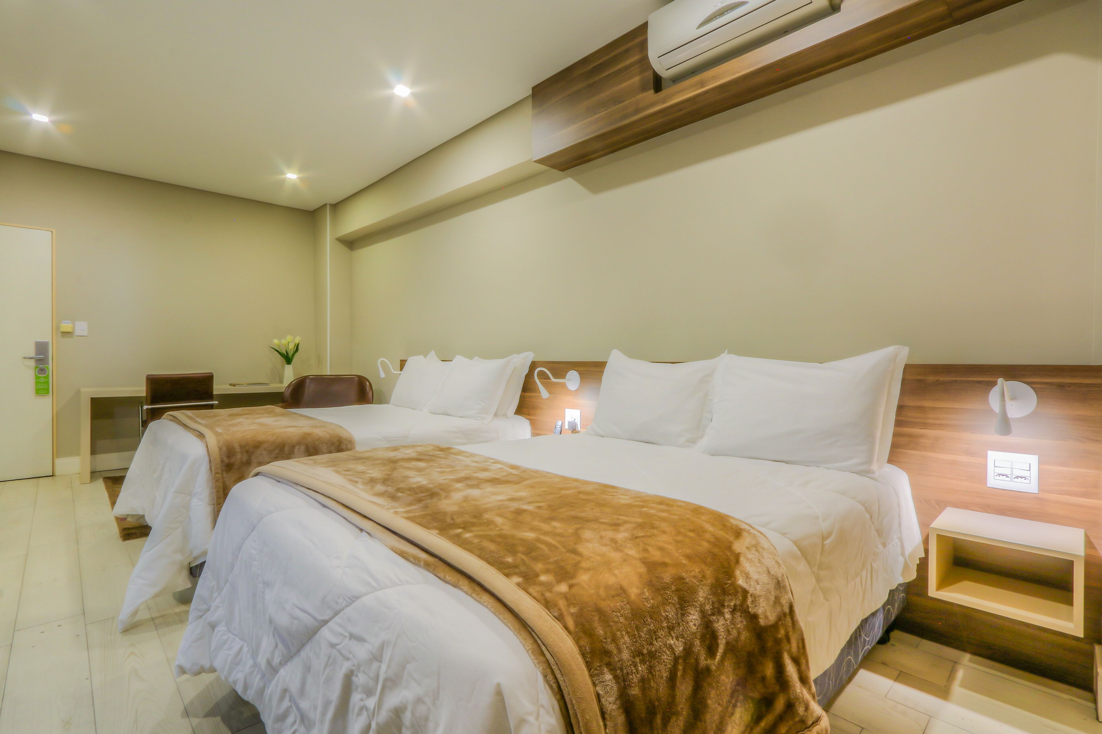 A hotel room with two beds, a desk, a chair, a nightstand, and an air conditioning unit.