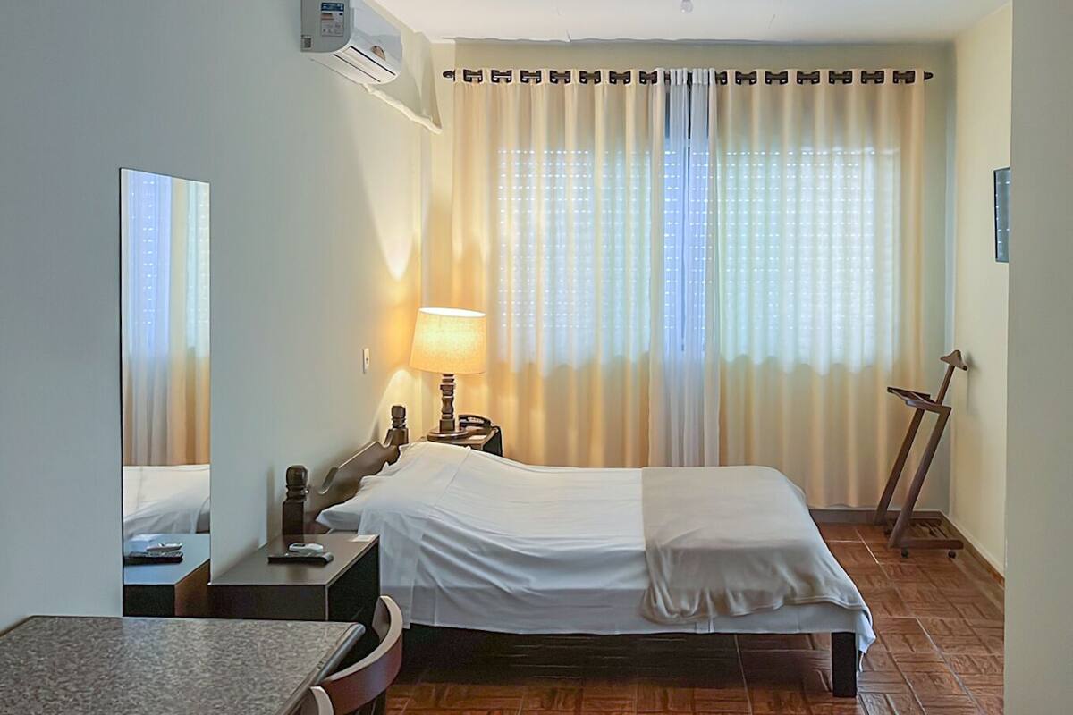 A hotel room with a bed, a desk, a chair, a lamp, a mirror, and a window with curtains.