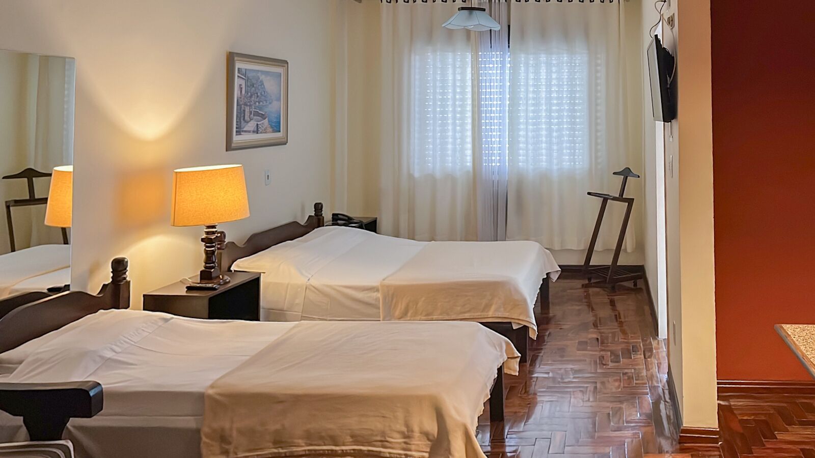 A hotel room with two beds, a nightstand with a lamp, a painting on the wall, and a window with sheer curtains.