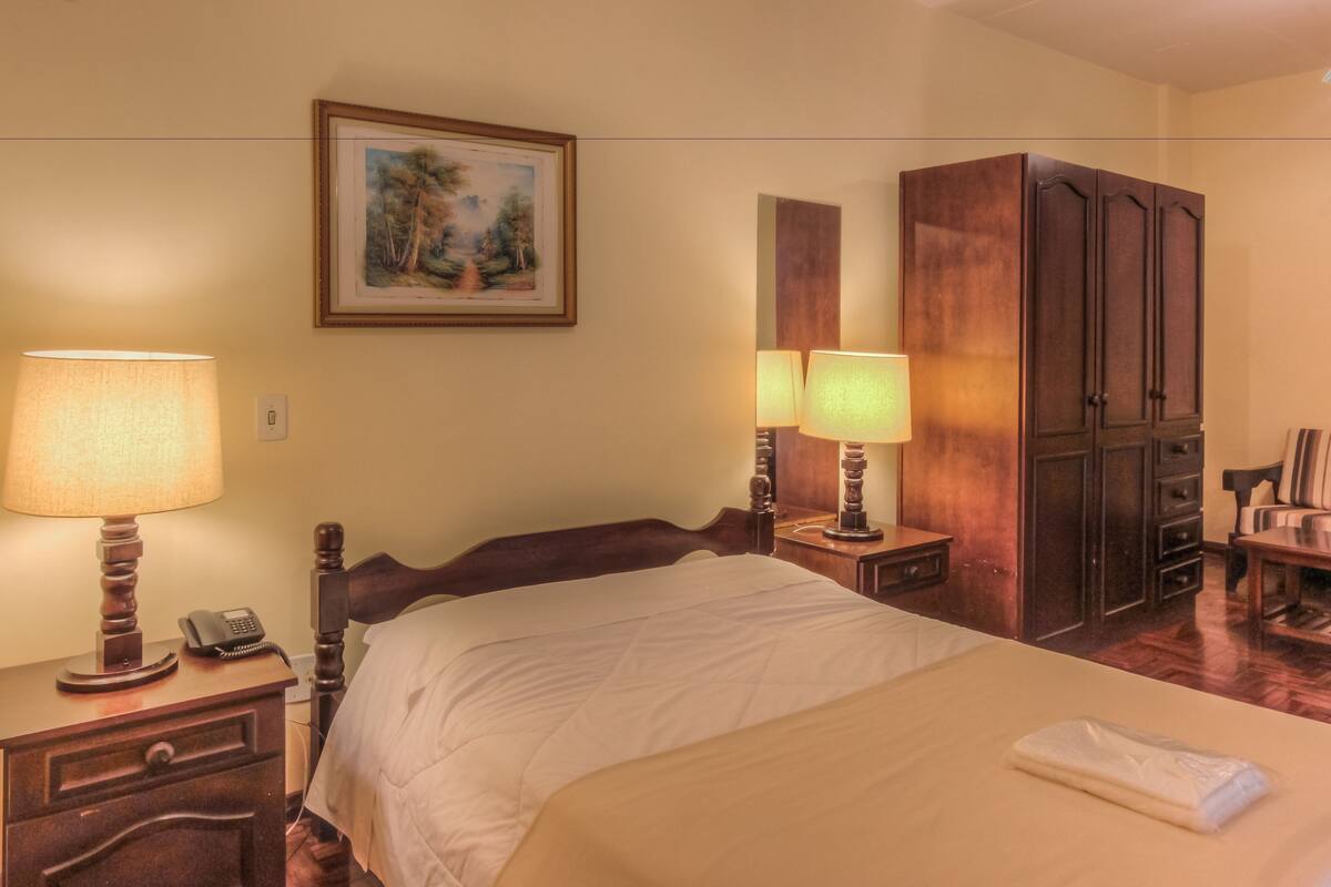 A hotel room with a bed, two bedside lamps, a telephone, a painting on the wall, and a wardrobe.