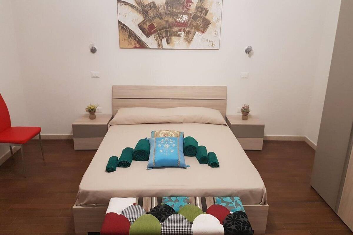 Een netjes opgemaakt bed met een beige hoofdbord, een klein nachtkastje met een plantje en een rode stoel links.