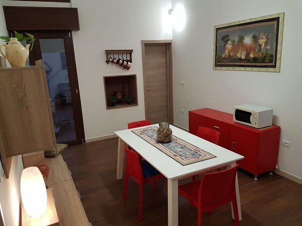 Un espacio habitable compacto con una mesa de comedor, sillas rojas, un mueble rojo con microondas, un estante pequeño con plantas y un cuadro enmarcado de una escena histórica.