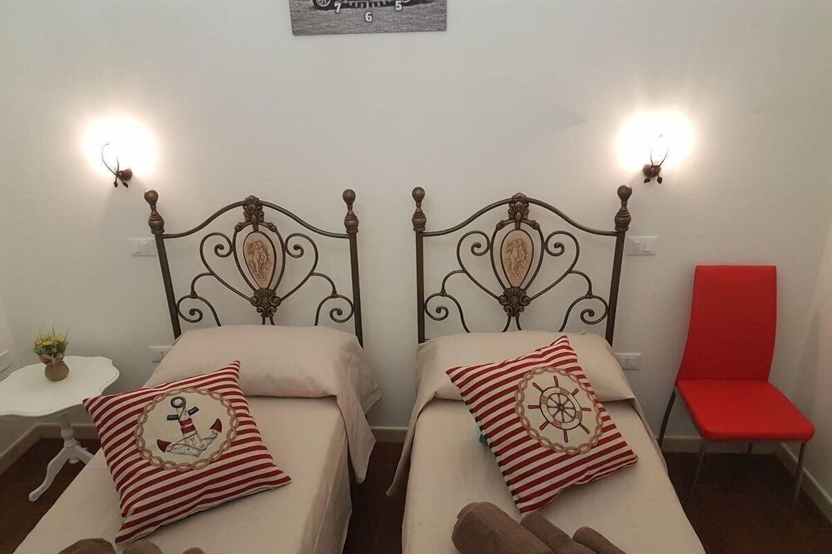 Twee enkele bedden met decoratieve metalen hoofdborden, elk met een wandlamp. Een klein tafeltje met een bloemenvaas en een rode stoel zijn zichtbaar.