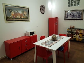 Un comedor con una mesa blanca, sillas rojas, un armario rojo con cajones, un microondas encima y un plato decorativo sobre la mesa.