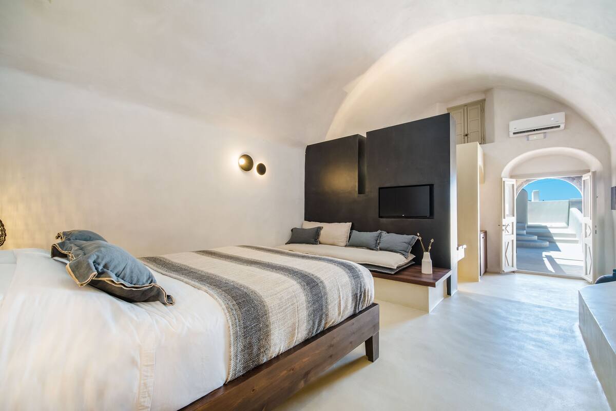 Une chambre à coucher comprenant un lit, des tables de chevet, un téléviseur fixé au mur et une porte en arc donnant sur un espace extérieur.