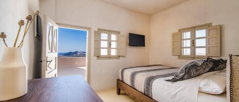 Un dormitorio con cama de madera, mesita de noche, televisor montado en la pared y ventana con contraventanas.