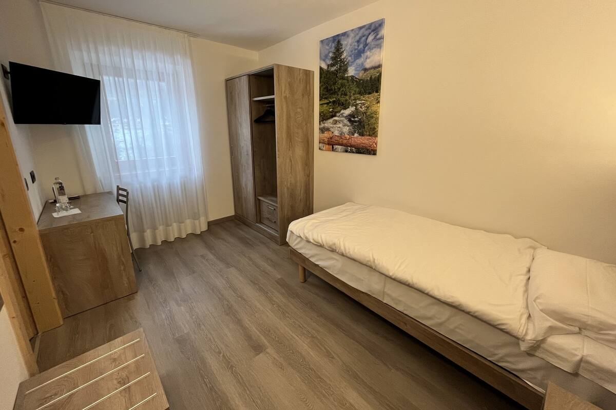 A bedroom with a bed, a desk with a chair, a wardrobe, and a TV mounted on the wall.