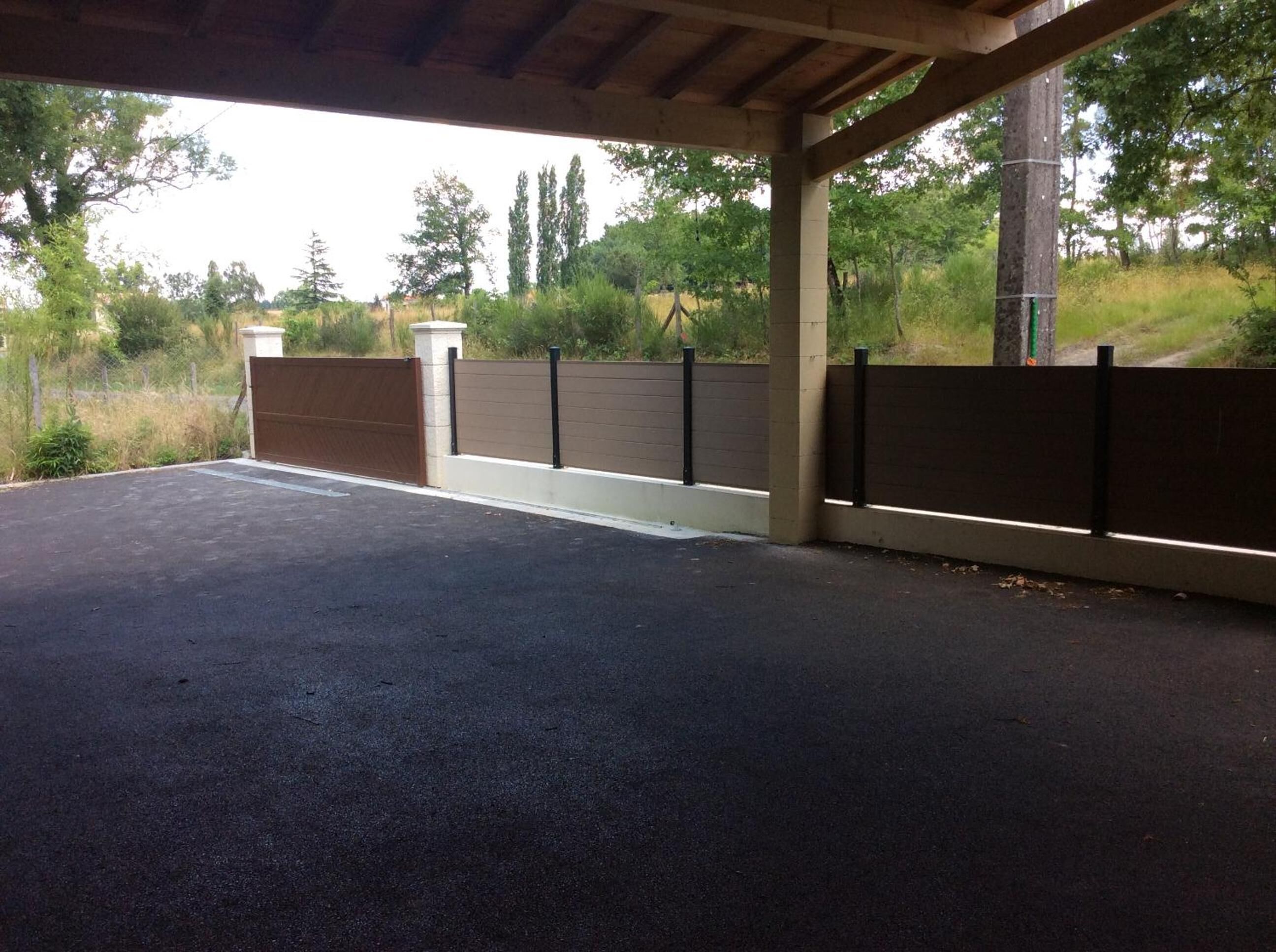 A covered parking area with a concrete floor, a wooden fence, and a view of trees and grass.