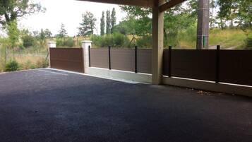 A covered parking area with a concrete floor, a wooden fence, and a view of trees and grass.