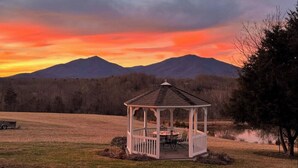 Un gazebo con tavolo e sedie in un prato erboso, circondato da montagne e un lago.