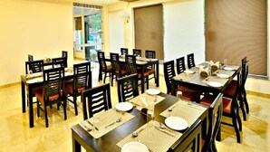 Comedor con varias mesas y sillas, dispuesto en una sala amplia con techo alto y ventanas grandes.