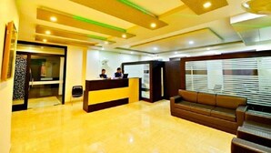 Zona de recepción de un hotel con mostrador, dos empleados, un sofá marrón y una escalera.