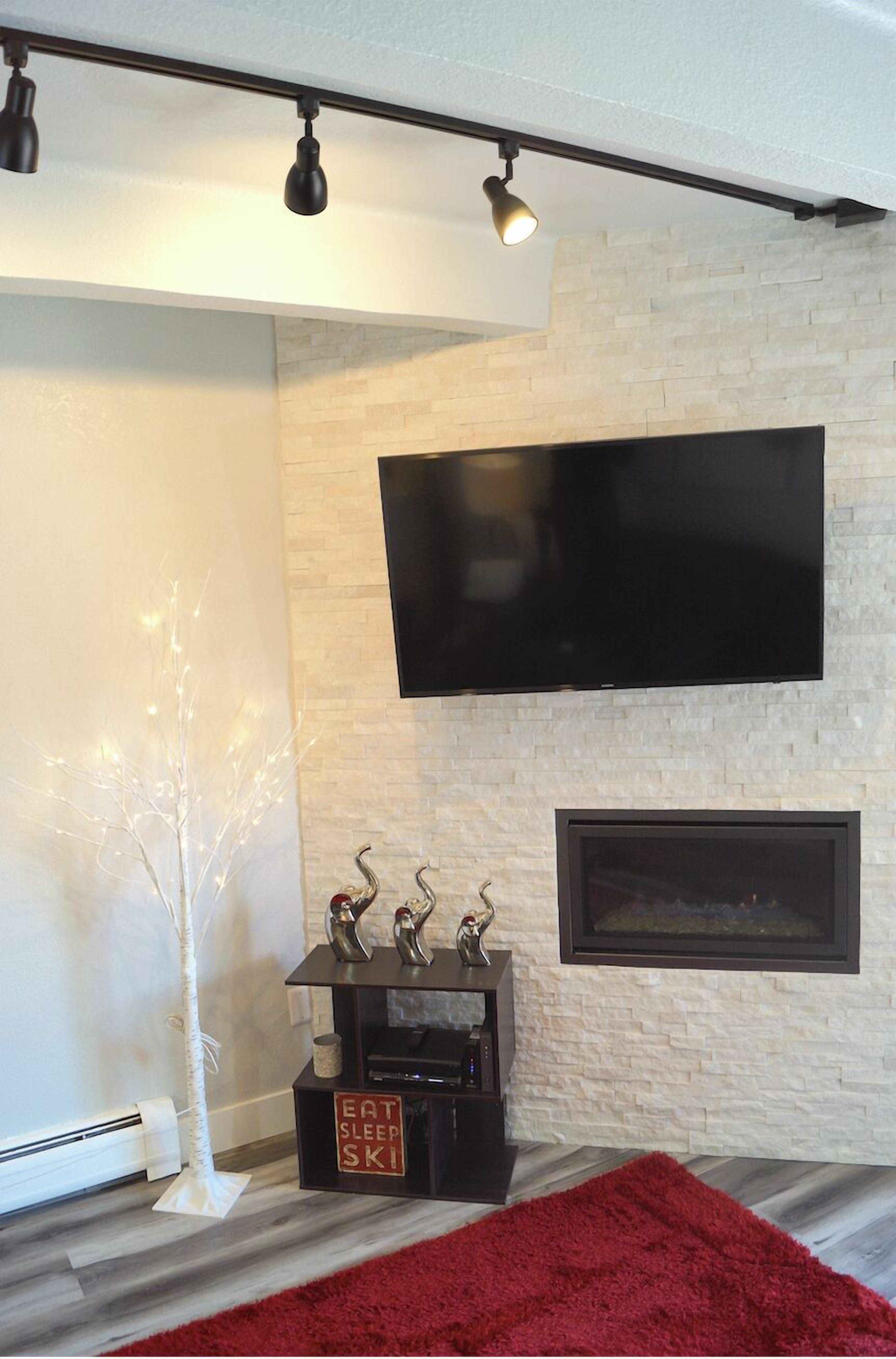 A modern living room with a flat-screen TV mounted on a white brick wall, a fireplace below, and a red rug on the floor.
