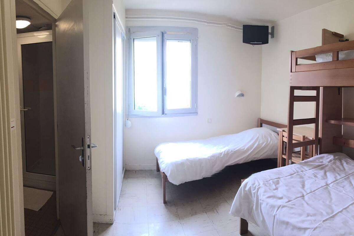 Een kamer met twee eenpersoonsbedden, een houten stapelbed, een raam met zonwering, een deur en een aan de muur bevestigde televisie.
