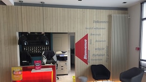 Un espace d’accueil avec un comptoir rouge, des chaises noires et un mur où sont affichés des messages de bienvenue dans plusieurs langues.