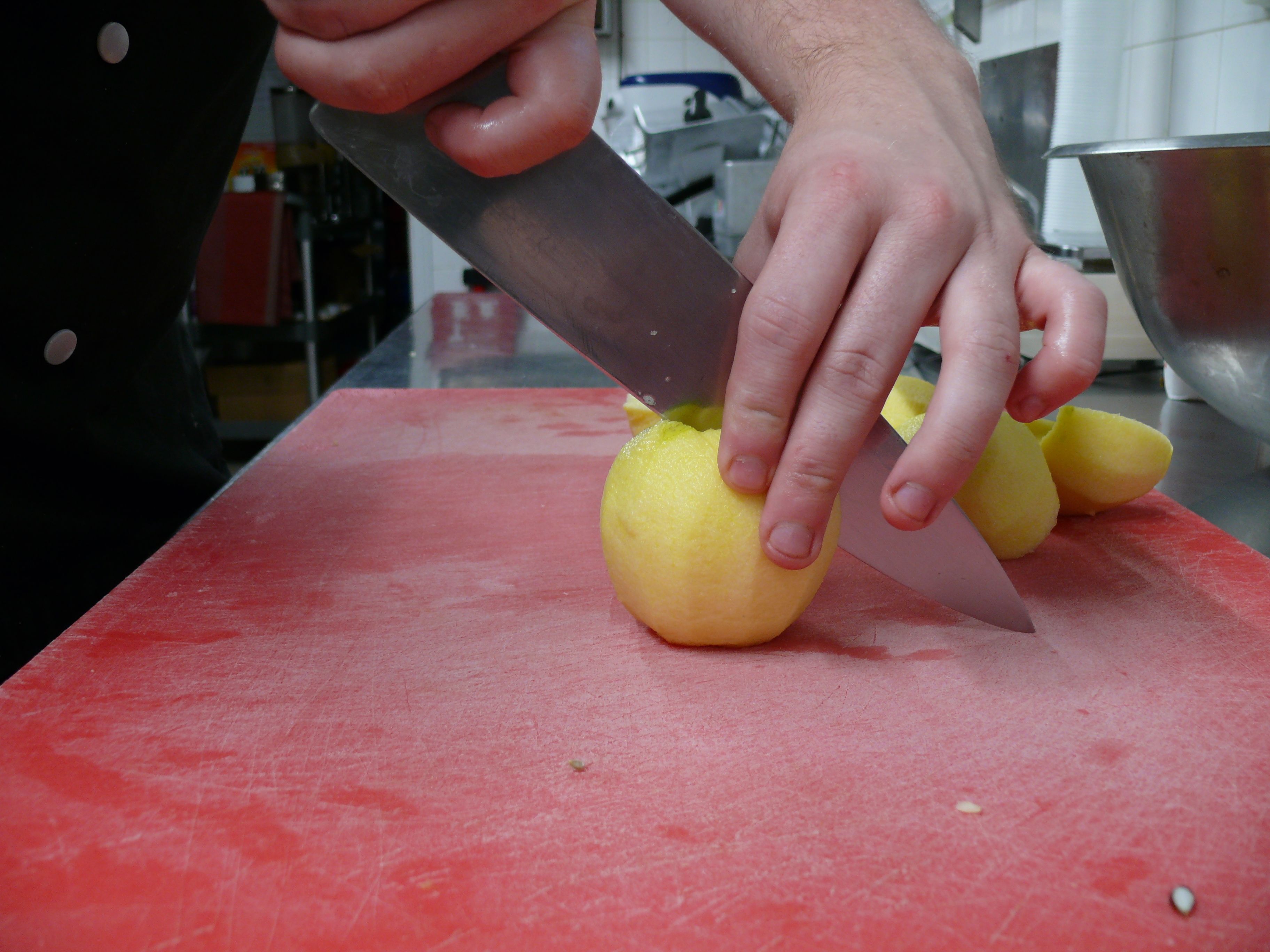 Una persona corta una fruta amarilla con un cuchillo grande sobre una tabla de cortar roja.