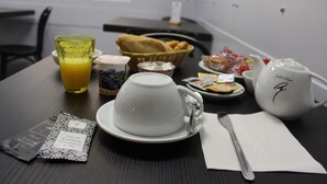 Una mesa de desayuno con una taza blanca, un vaso de zumo de naranja, una cesta de cruasanes, un plato de pasteles, una tetera, una bolsita de té y una bolsita de café.
