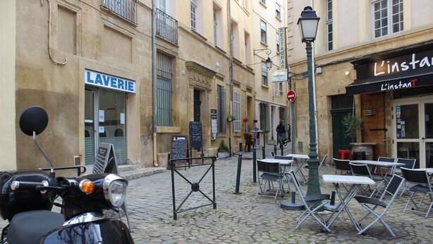 Una calle empedrada con una moto estacionada, zona de mesas y sillas al aire libre, y locales comerciales que incluyen una lavandería y un restaurante.