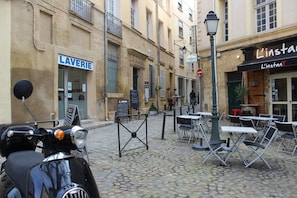 A cobblestone street with a parked scooter, outdoor seating area with tables and chairs, and storefronts including a laundry service and a restaurant.