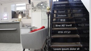 Un mostrador de recepción con una computadora, una silla con cojín rojo y una escalera con texto hebreo.