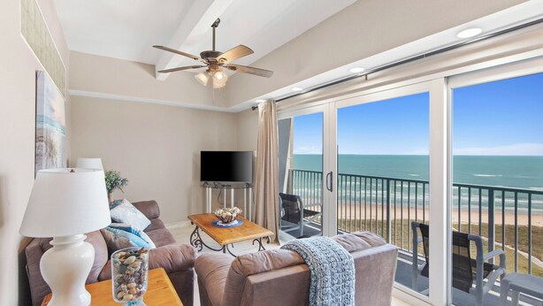 Un balcon avec vue sur l’océan, un ventilateur de plafond, un téléviseur à écran plat et un coin salon avec une table et des coussins.