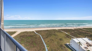 Une vue sur la plage depuis un balcon, avec un ciel dégagé, une plage de sable et un bâtiment blanc orné d’un palmier.