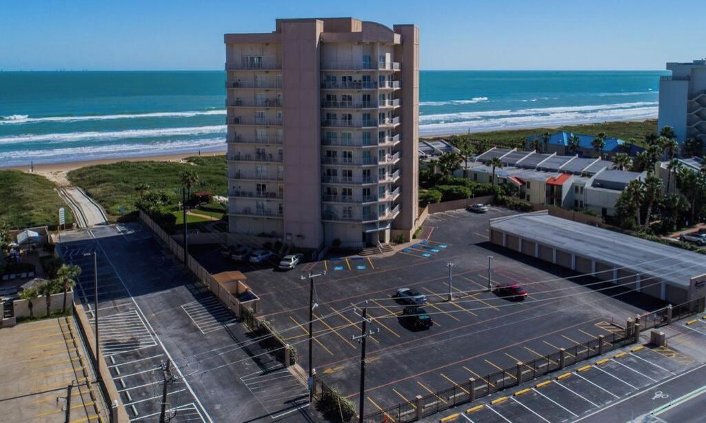 Un hotel de varios pisos con estacionamiento frente a él, vista al mar y cielo despejado.