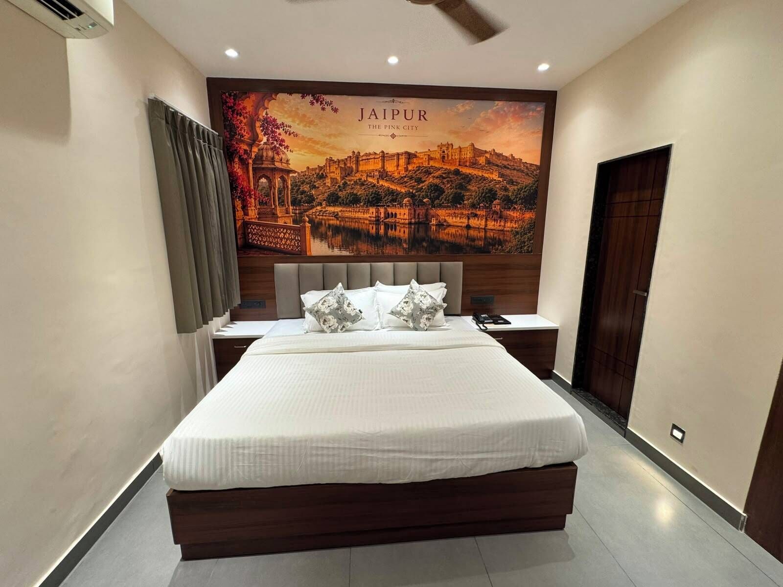 Habitación de hotel con una cama grande, una mesita de noche, aire acondicionado de pared y un cuadro enmarco de Jaipur.