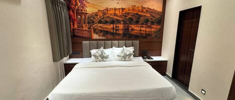 A hotel room with a large bed, a nightstand, a wall-mounted air conditioner, and a large framed picture of Jaipur.