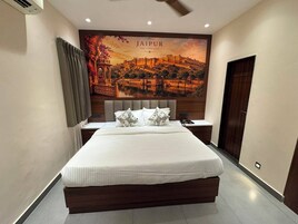 A hotel room with a large bed, a nightstand, a wall-mounted air conditioner, and a large framed picture of Jaipur.