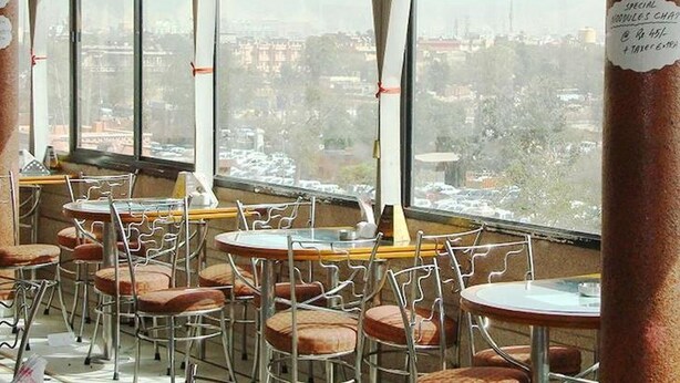 Un restaurante con varias mesas y sillas, amplios ventanales que ofrecen vistas a la ciudad y un letrero con los precios.