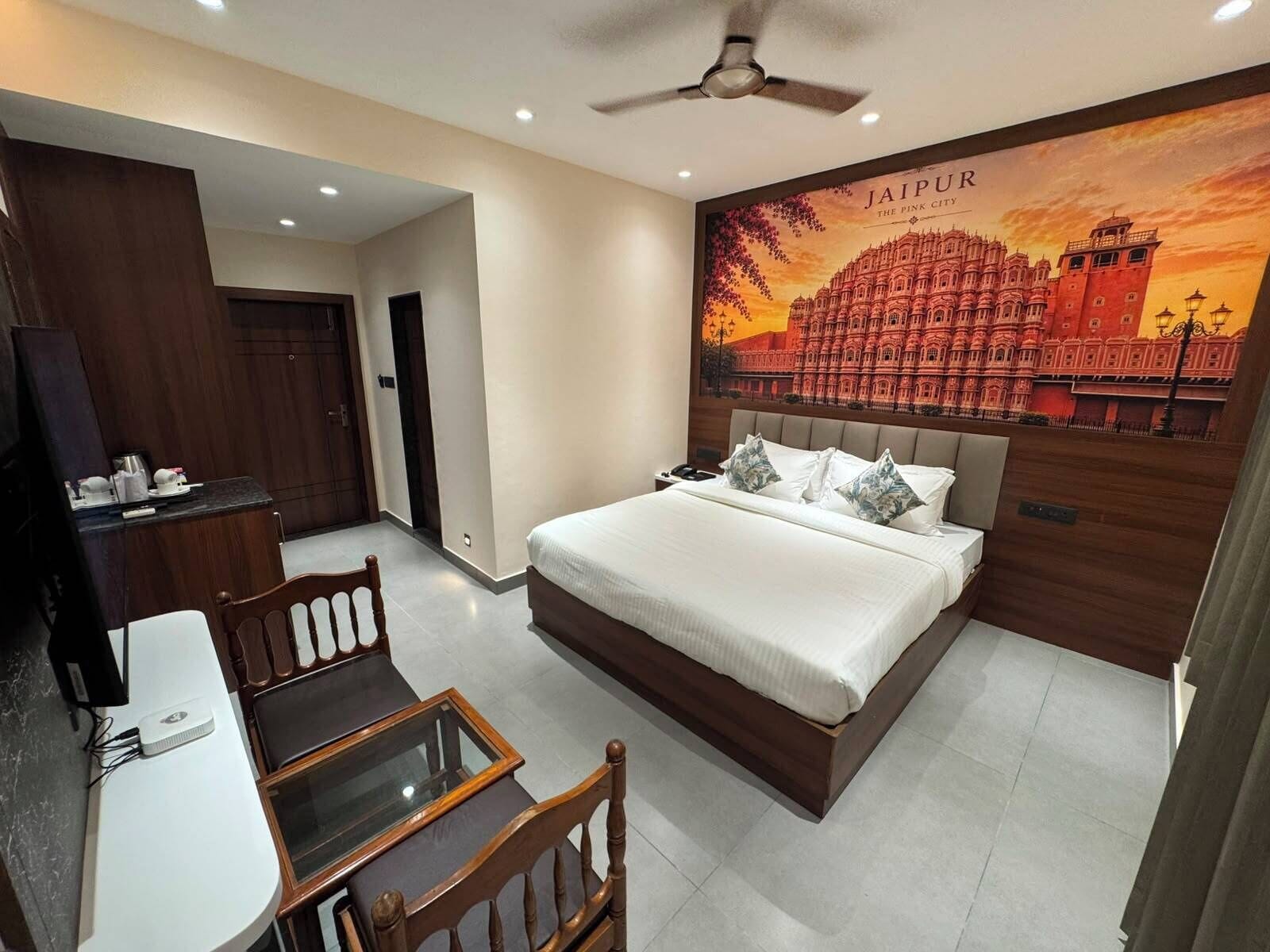 Hotelkamer met een bed, een bureau, een stoel, een plafondventilator en een wanddecoratie van Jaipur.