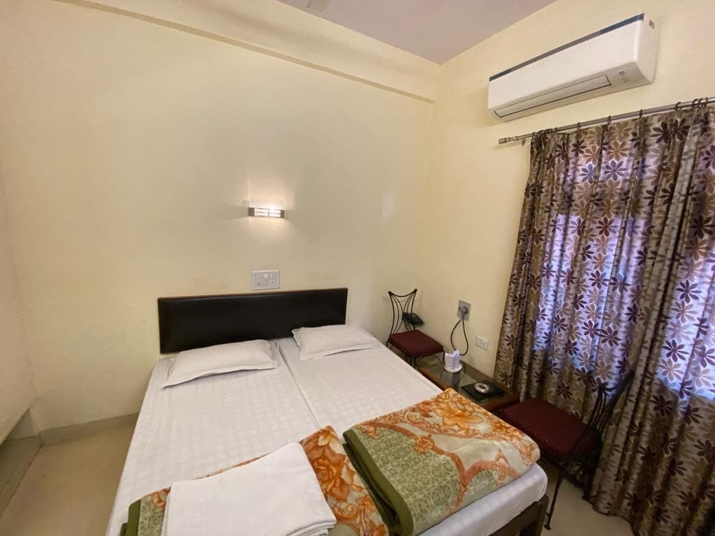 A hotel room with a bed, a nightstand, an air conditioning unit, and a window with curtains.