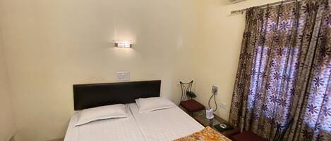 A hotel room with a bed, a nightstand, an air conditioning unit, and a window with curtains.