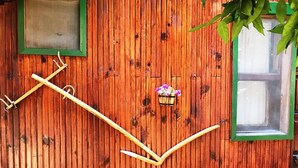 A wooden wall with a green-framed window and a small window. There is a wooden structure resembling a book or a letter 'L' and a small basket with flowers. - Divasa Hotel (Kumluca)