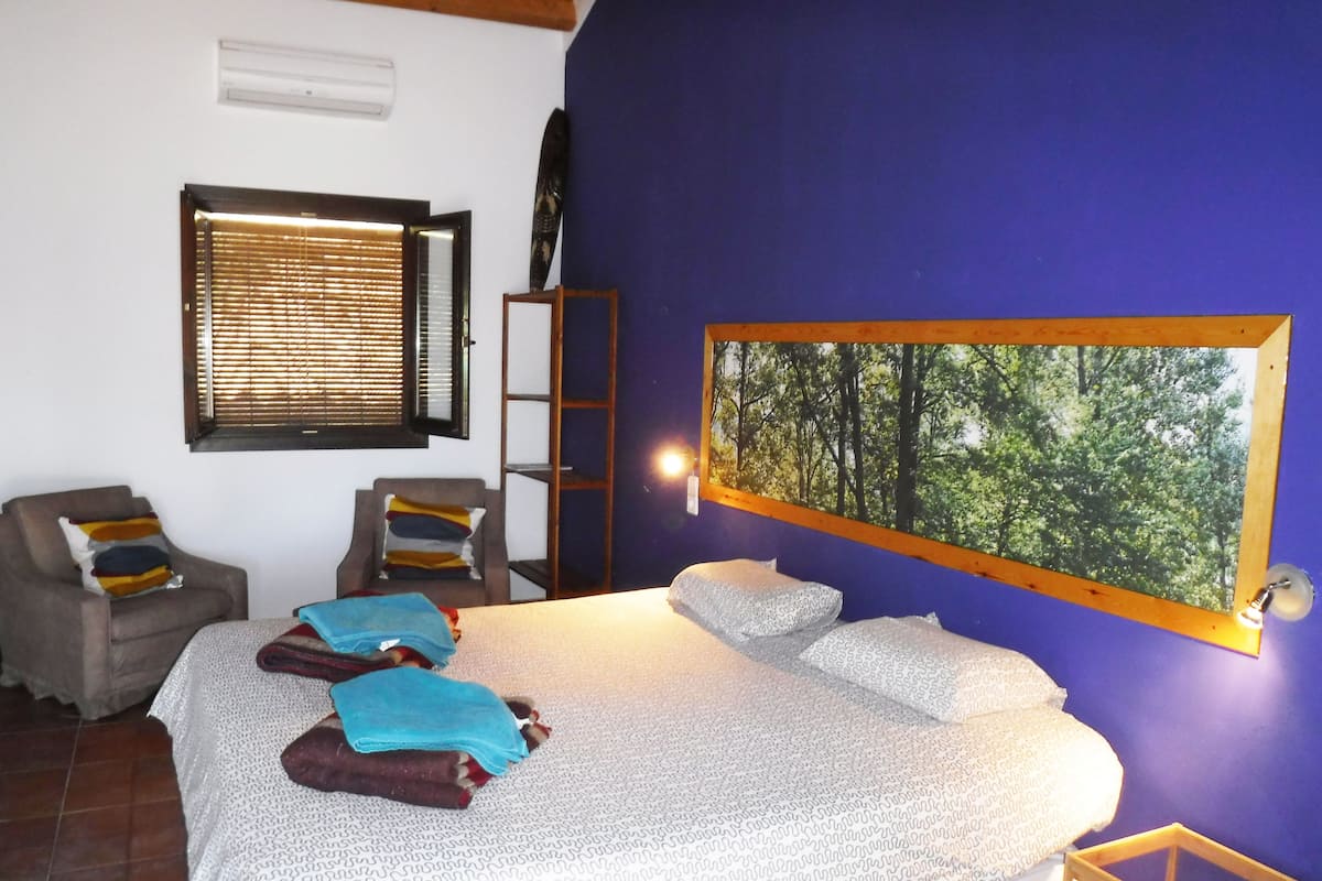 Ein Schlafzimmer mit einem Bett, zwei Stühlen, einer Leiter, einem Fenster mit Jalousien und einem an der Wand hängenden Bild von einem Wald.