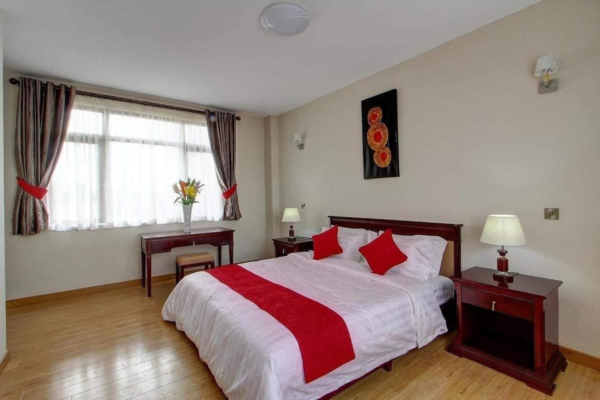A hotel room with a large bed, two bedside tables with lamps, a desk with a chair, a window with curtains, and a wall-mounted artwork.
