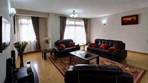 A living room with two black leather sofas, a coffee table, a TV, and a large window with curtains. - Fenesi Gardens Apartments (Nairobi)