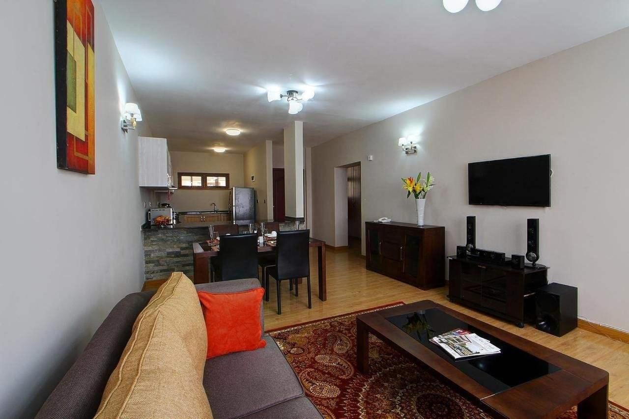 Un salon moderne avec un canapé, une table basse et un téléviseur à écran plat. On y trouve également un coin repas avec des chaises et une table, et une cuisine à l’arrière-plan.