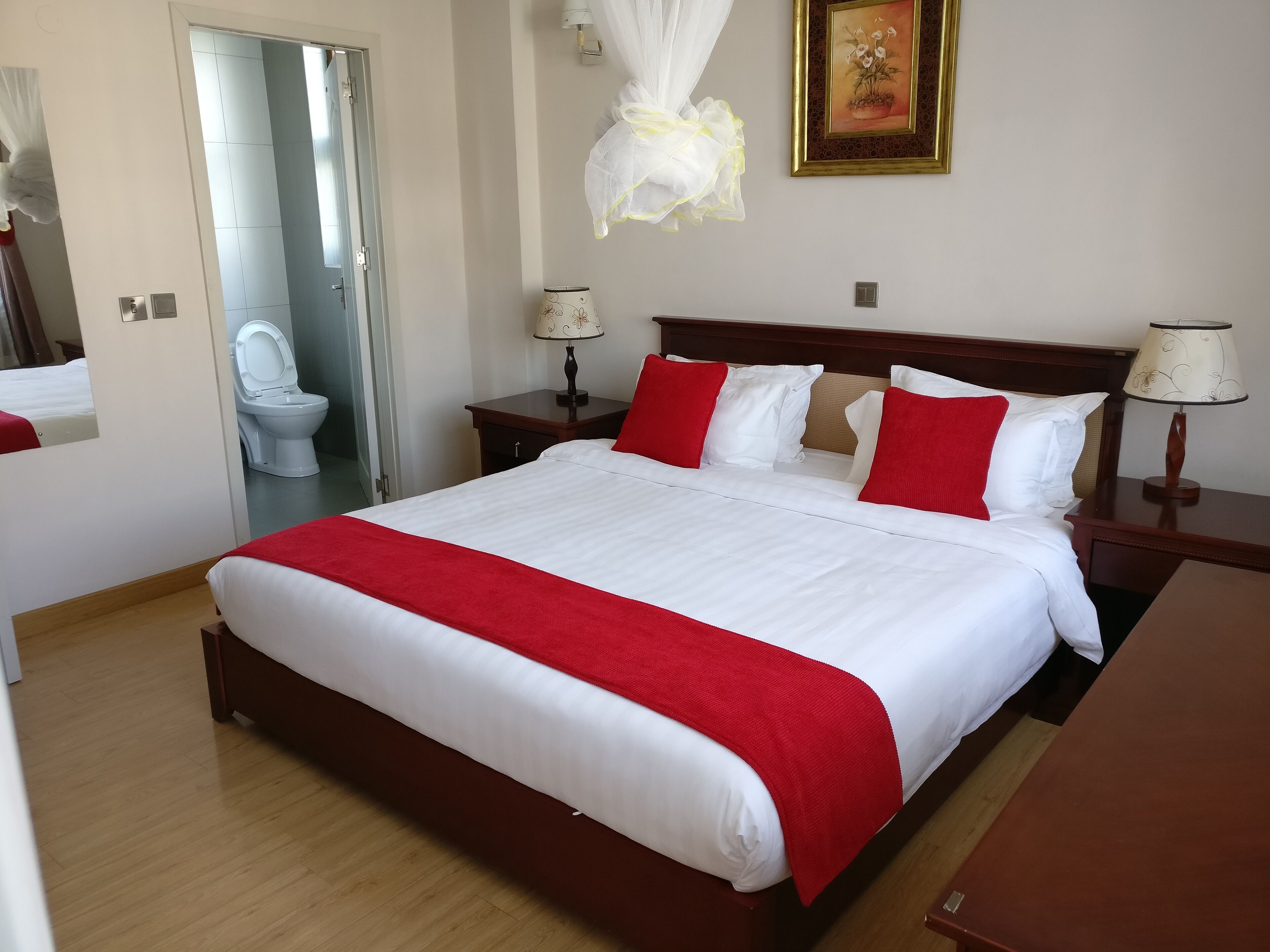 A neatly made bed with a red and white bedspread, two bedside tables with lamps, a bedside mirror, a painting on the wall, and a door leading to a bathroom.