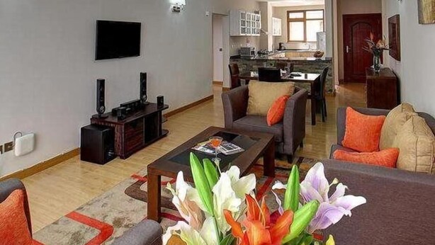 A modern living room with a flat-screen TV, a coffee table, and a sofa. There is a kitchen in the background with cabinets and a dining area. - Fenesi Gardens Apartments (Nairobi)